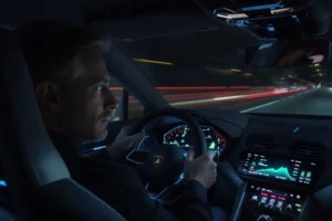 Lamborghini aposta em inteligência artificial para detectar estado emocional do motorista Close-up de um homem dirigindo um Lamborghini à noite, com painel digital exibindo indicador de emoção.