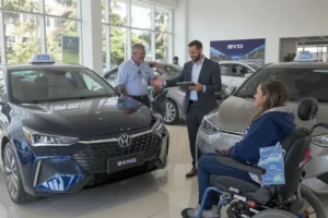 BYD King e Song Pro têm descontos de até R$ 57 mil para taxistas e PCD Um taxista e uma mulher em cadeira de rodas, ambos sorrindo, em uma concessionária BYD, observando os modelos King e Song Pro, com um vendedor explicando os benefícios.